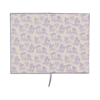 An open book reveals patterned endpapers featuring a repeating light purple floral and pastoral scene on a cream background, with a lavender ribbon bookmark draped down the center. The Very Tired Woman Journal showcases a bookcloth cover and lay-flat binding for effortless writing or sketching across its pages.