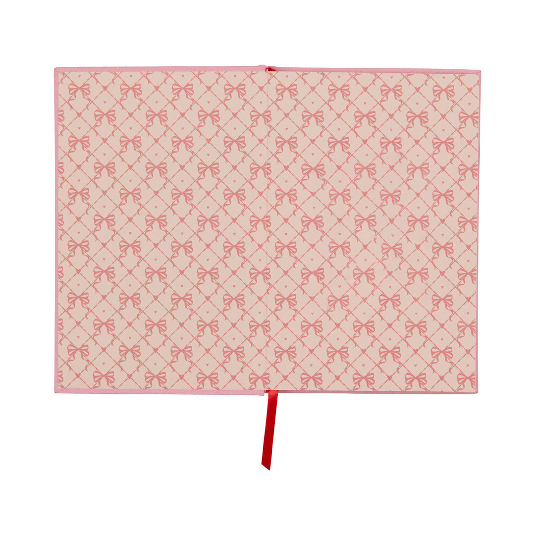 A flat, pink rectangular “It’s All Going Swell Journal” features a vintage-inspired design with a repeated pattern of red bows and diamonds, accented by a single red ribbon extending from the center of the bottom edge.
