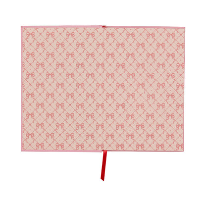 A flat, pink rectangular “It’s All Going Swell Journal” features a vintage-inspired design with a repeated pattern of red bows and diamonds, accented by a single red ribbon extending from the center of the bottom edge.