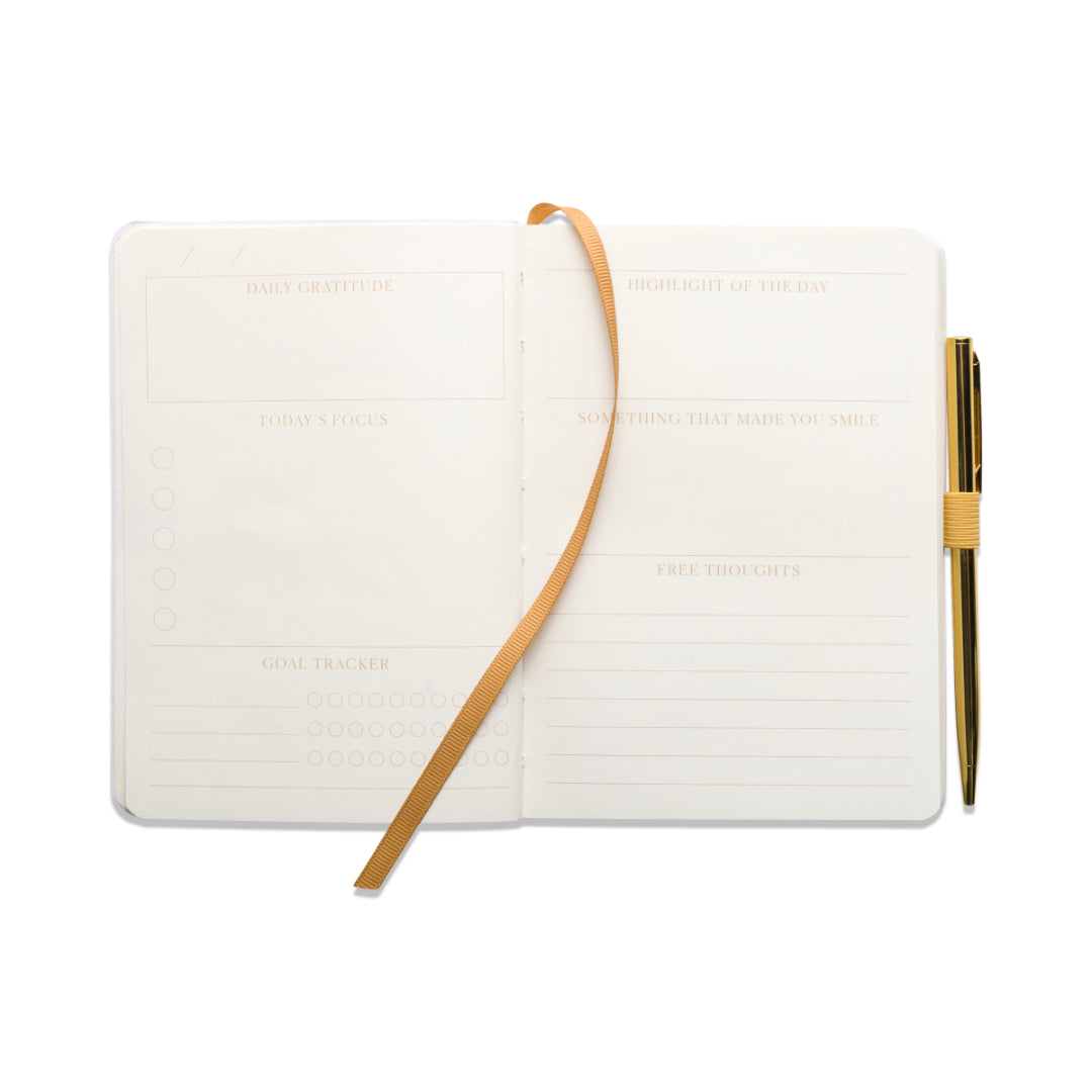The Gratitude Journal with Pen - You Are Exactly Where You Need To Be features guided sections for gratitude, today’s focus, goals, highlights, something that made you smile, and free thoughts. It comes with a gold pen and a ribbon bookmark attached.
