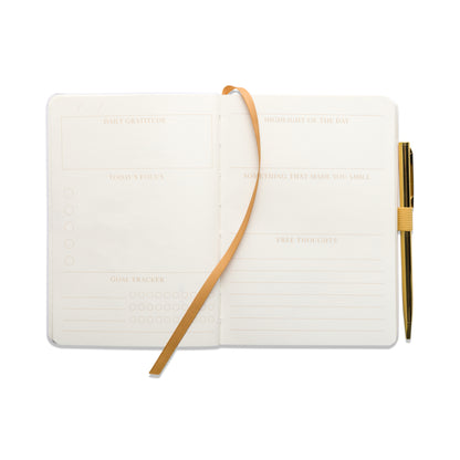 The Gratitude Journal with Pen - You Are Exactly Where You Need To Be features guided sections for gratitude, today’s focus, goals, highlights, something that made you smile, and free thoughts. It comes with a gold pen and a ribbon bookmark attached.