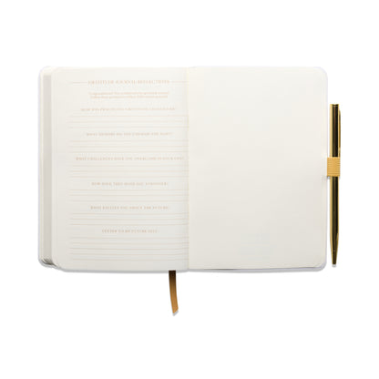 The “Gratitude Journal with Pen – You Are Exactly Where You Need To Be” features guided prompts on the left side of each spread and lined pages for writing on the right, accompanied by a gold pen secured in a loop and a yellow ribbon bookmark, all set against a white background.