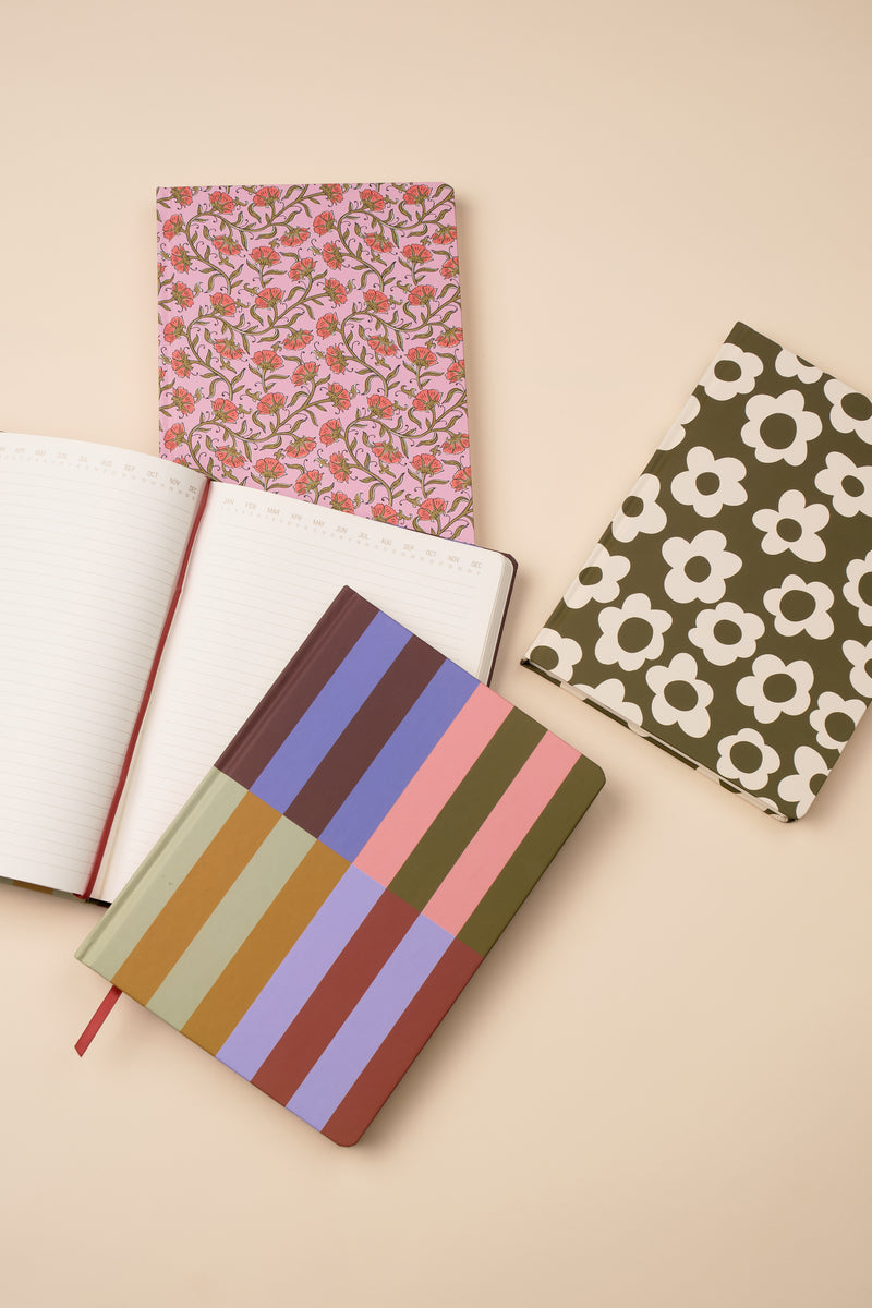 Four notebooks rest on a beige surface. Among them is the Soft Touch Jumbo Journal - Bold Stripe, an oversized notebook featuring a bold striped cover. One notebook is open, displaying its neatly lined pages.