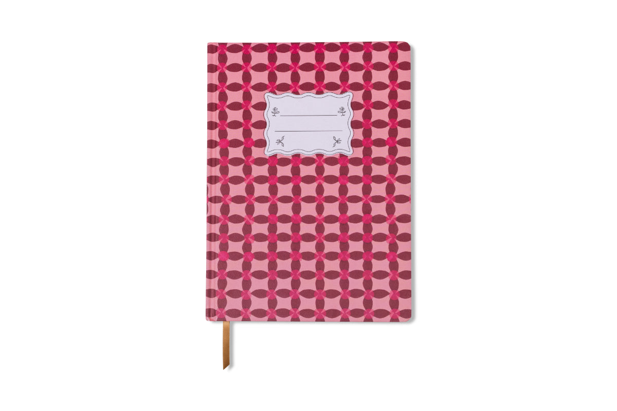 The Tulip Jumbo Journal - Pinwheel Pink features a repeating red geometric pattern, a white front label, and a brown ribbon bookmark. This floral-inspired journal stands out on a crisp white background.