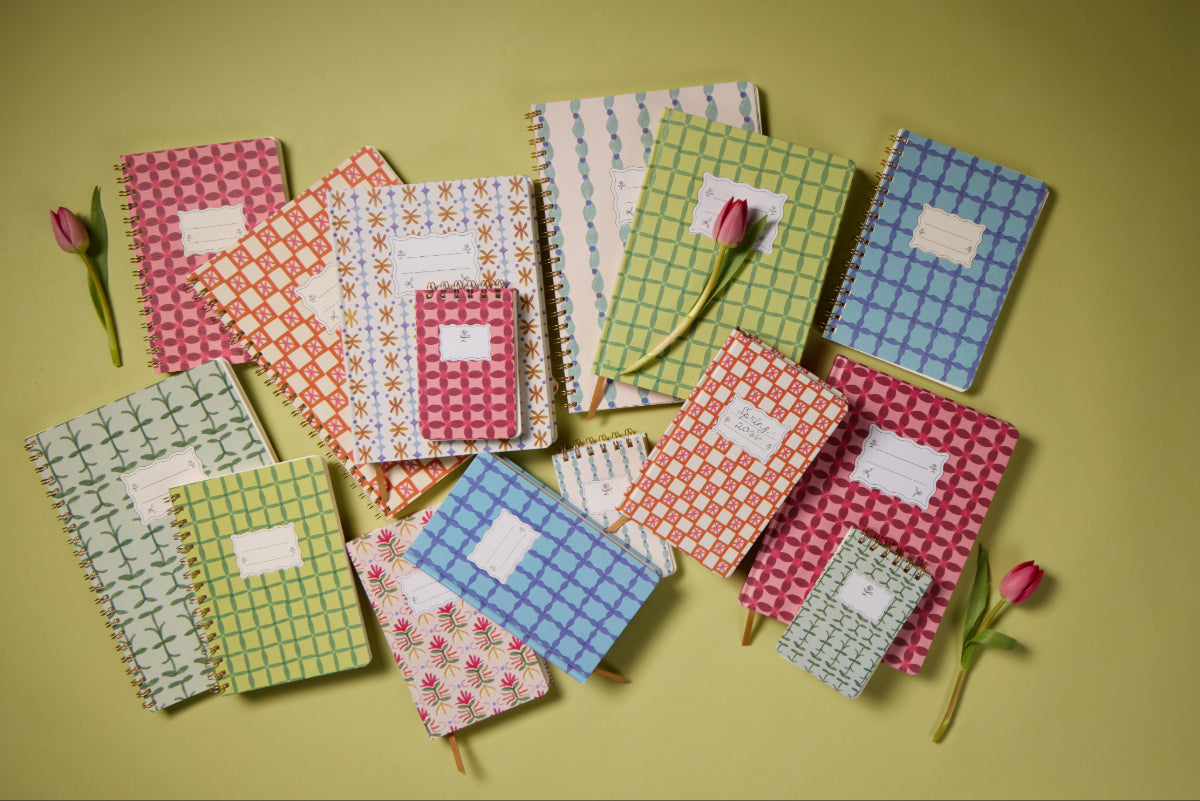 The Tulip Twin Wire Notebook - Large Green Vine and other colorful, patterned notebooks with blank labels are arranged on a green surface, accompanied by three small pink tulips.