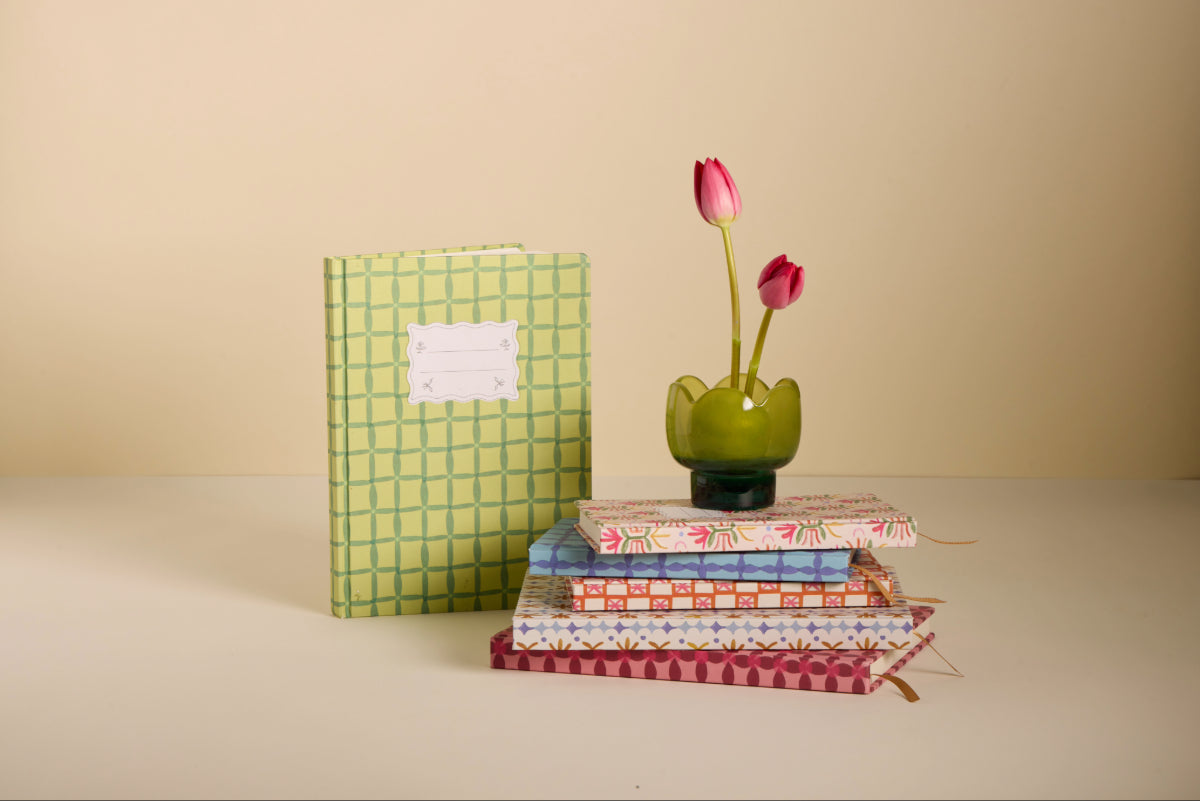 A green Tulip Jumbo Journal - Green Lattice stands upright next to a small stack of patterned journals. Nearby, a green vase with two pink tulips sits against a beige background.