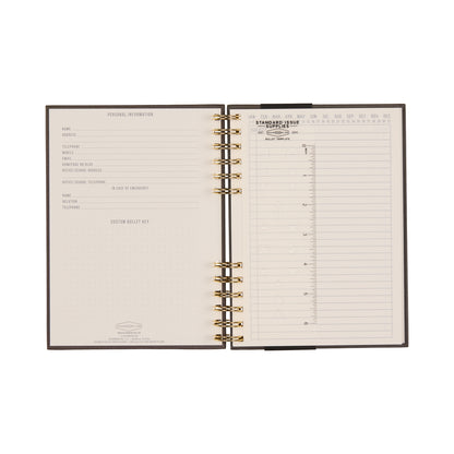 An open Standard Issue Notebook No.12 - Black, featuring high-quality construction, displays a personal information form on the left and a lined undated monthly calendar with columns for each day of the month on the right, offering a premium writing experience reminiscent of the renowned Standard Issue collection.