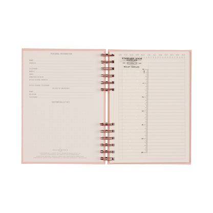 Introducing the Standard Issue Planner Notebook No. 12 - Rosewood + Blush: an open spiral-bound planner featuring a pink cover and a clean, organized layout inspired by the classic Standard Issue Notebook. Includes dedicated pages for personal information, a customizable bullet key, and versatile bullet journal templates such as monthly and date sections alongside dot grid pages—perfect for a premium writing experience.