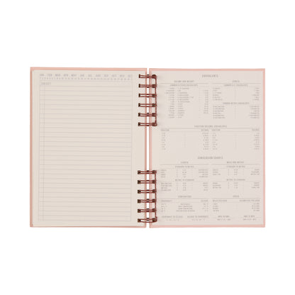The Standard Issue Planner Notebook No. 12 - Rosewood + Blush is an open spiral-bound notebook displaying a blank table on the left and assorted measurement and conversion charts on the right. Featuring a light pink cover and crisp white pages with printed text, this premium planner ensures a superior writing experience with its high-quality construction.