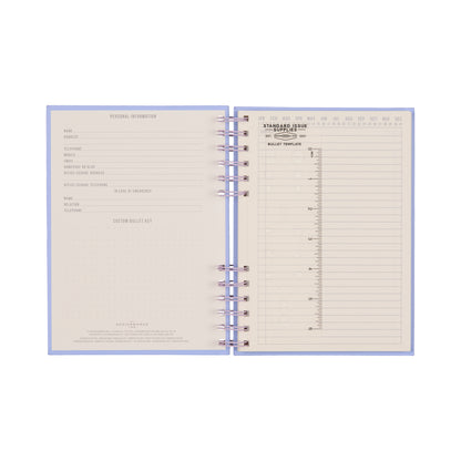 The Standard Issue Planner Notebook No. 12 - Lavender + Periwinkle features an open spiral-bound design, displaying a personal information page on the left and a yearly overview with monthly layouts and a bullet journal template on the right. Its cream-colored pages with gray text and dotted grids provide a premium writing experience.