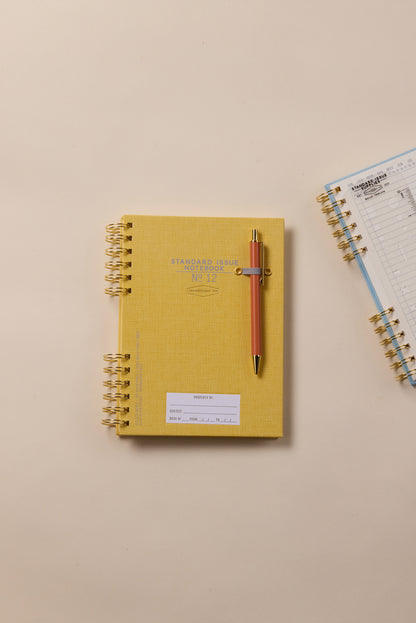 The Standard Issue Notebook No.12 - Ochre, paired with a gold and peach pen, rests on a beige surface for a premium writing experience. To its right, an open spiral-bound notebook lies nearby.