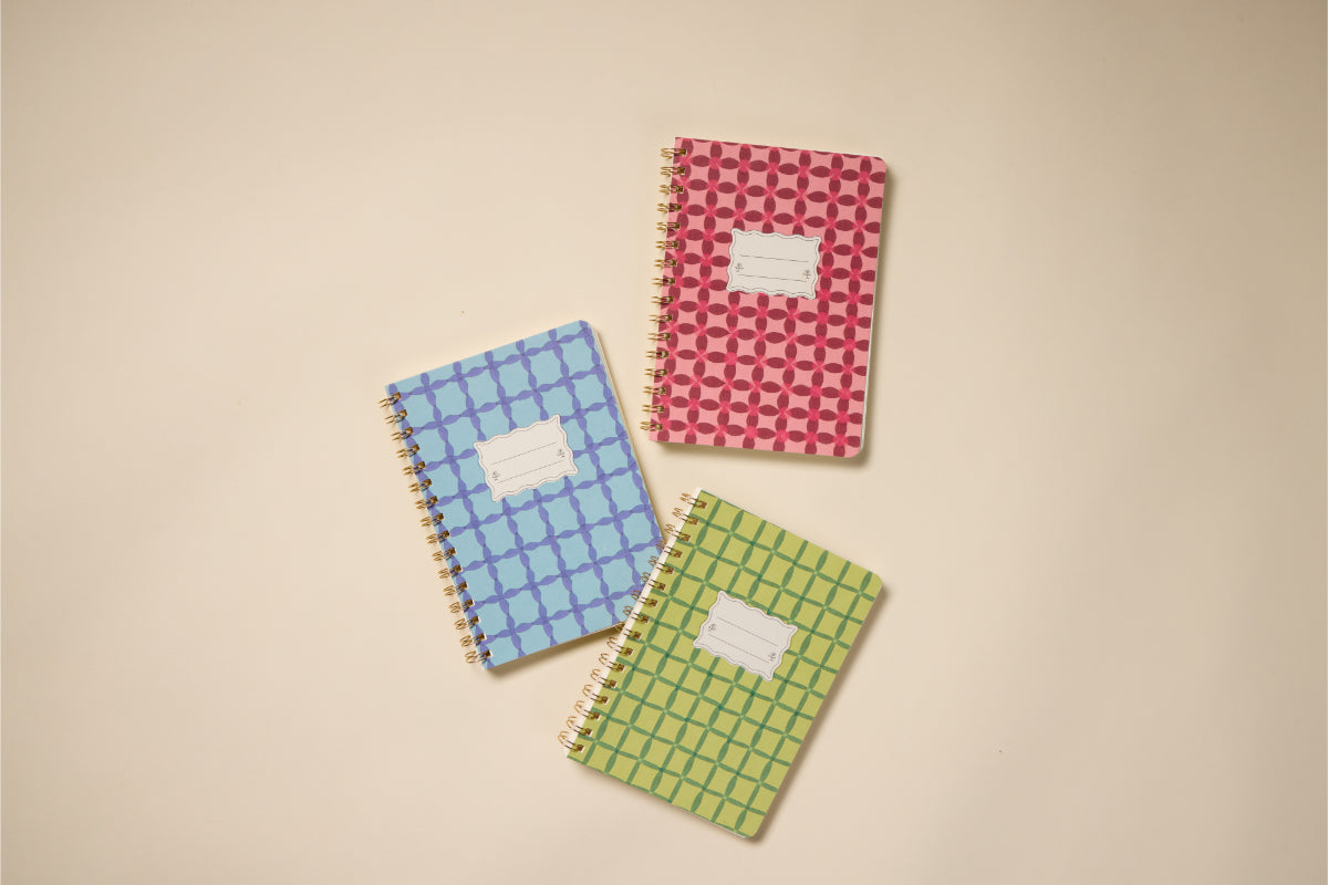 A Tulip Twin Wire Notebook - Small Green Lattice, along with blue and pink patterned spiral notebooks, lies on a beige surface. Each features a white label area on the front.