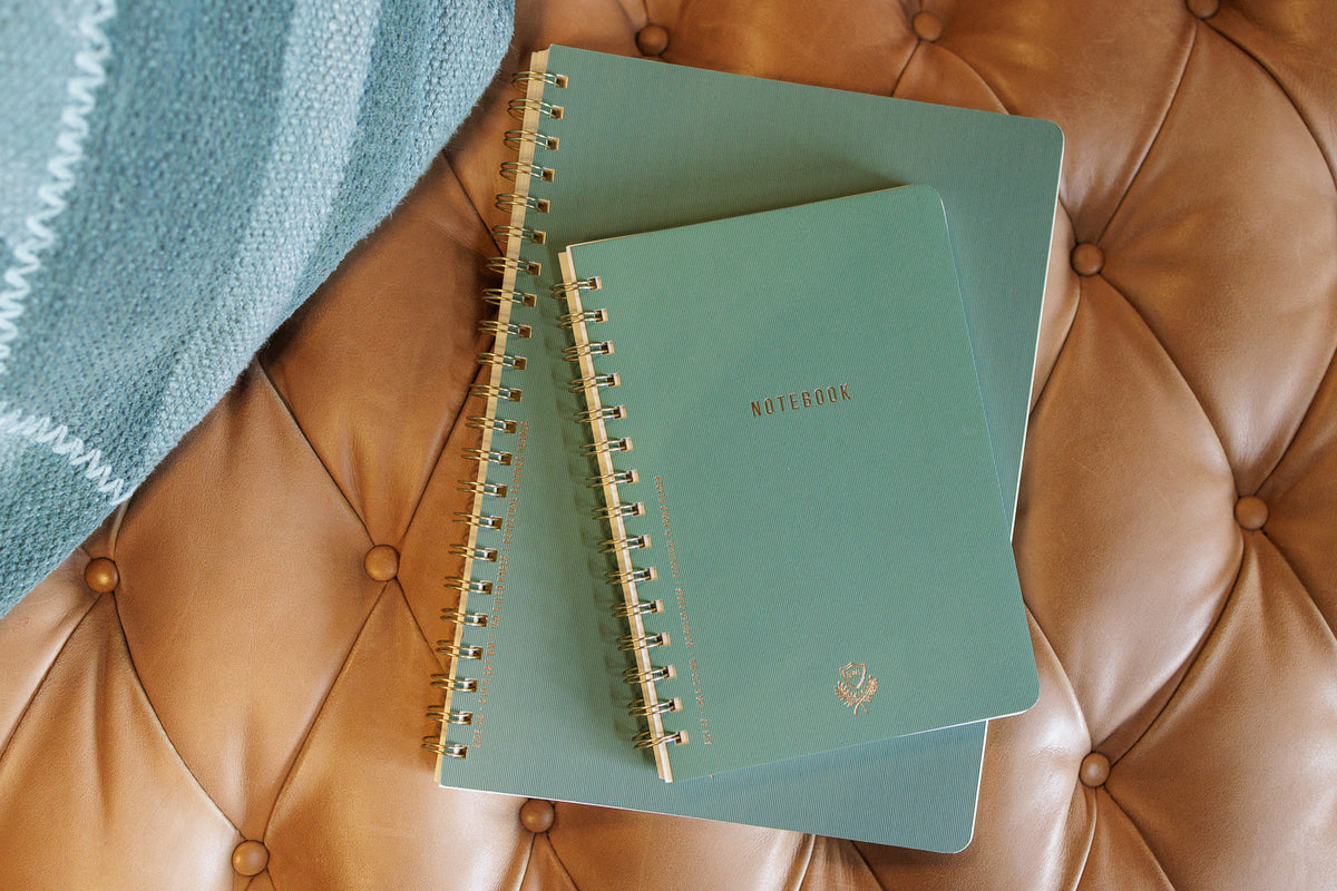 Two Textured Paper Twin Wire Notebooks in Large Juniper are stacked on a tan tufted leather surface beside a folded green patterned blanket.