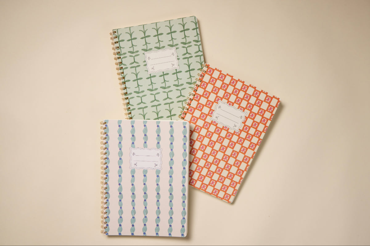 Three spiral-bound notebooks—including the Tulip Large Twin Wire Notebook - Green Vine with leaf designs, a red-orange geometric notebook, and a white botanical notebook with blue and teal lines—are arranged on a light beige surface.