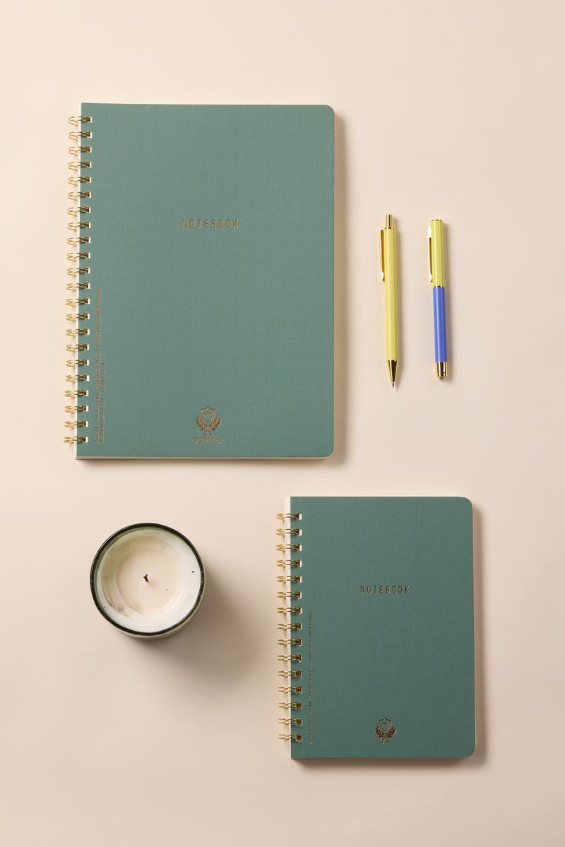A Textured Paper Twin Wire Notebook - Small Juniper rests on a light beige surface with a large green notebook, a white candle in a glass jar, and two pens, one yellow and one blue.