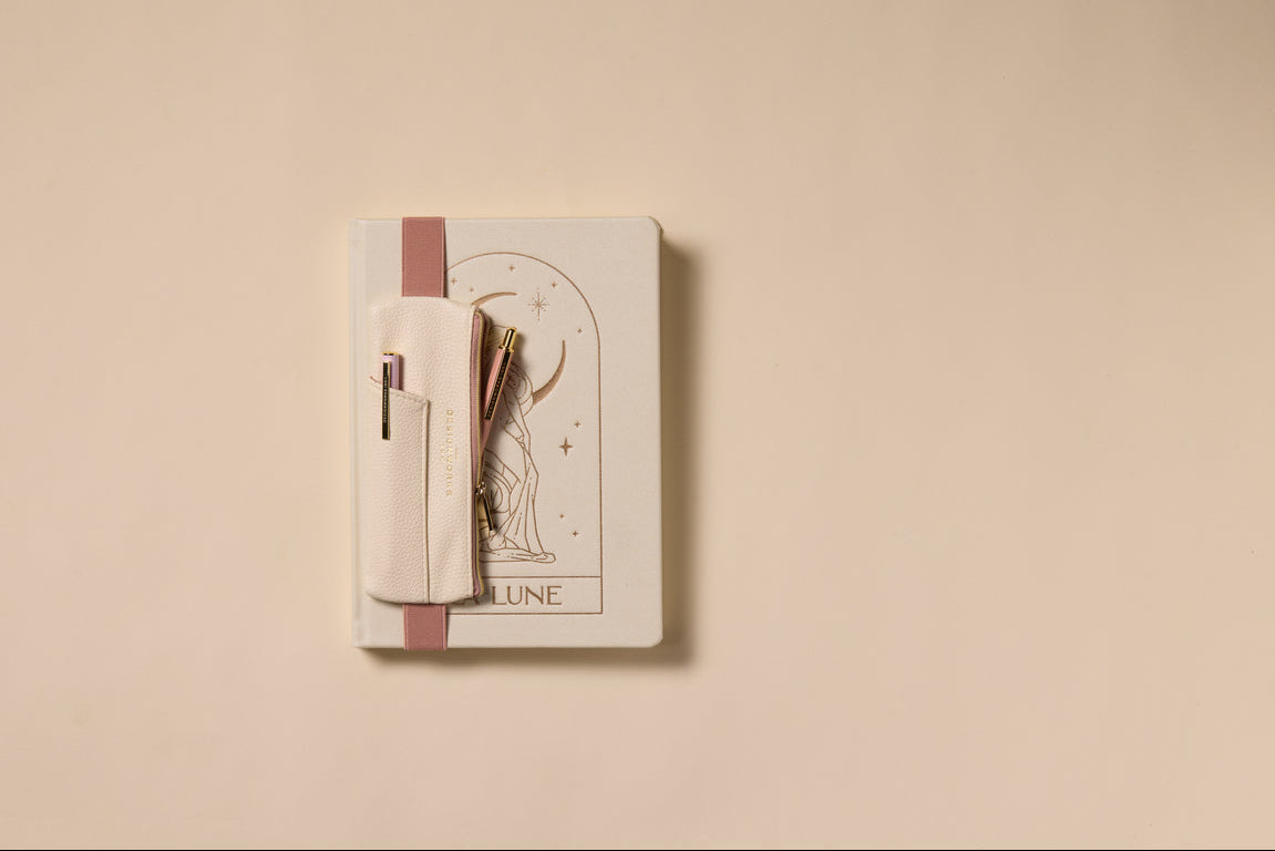 A closed beige notebook with a cat drawing and "LUNE" on the cover sits on a light surface. A Vegan Leather Pen Pouch in Cream + Taupe holds pens securely, attached to the notebook.