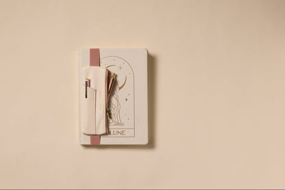 A closed beige notebook with a cat drawing and "LUNE" on the cover sits on a light surface. A Vegan Leather Pen Pouch in Cream + Taupe holds pens securely, attached to the notebook.