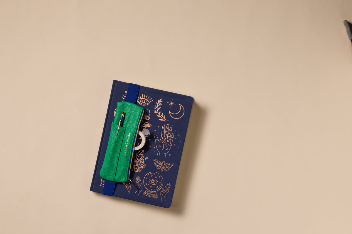A closed blue journal with gold mystical designs features a Vegan Leather Pen Pouch in bright green and navy, complete with a pen, all secured by an elastic band and placed on a plain beige surface.