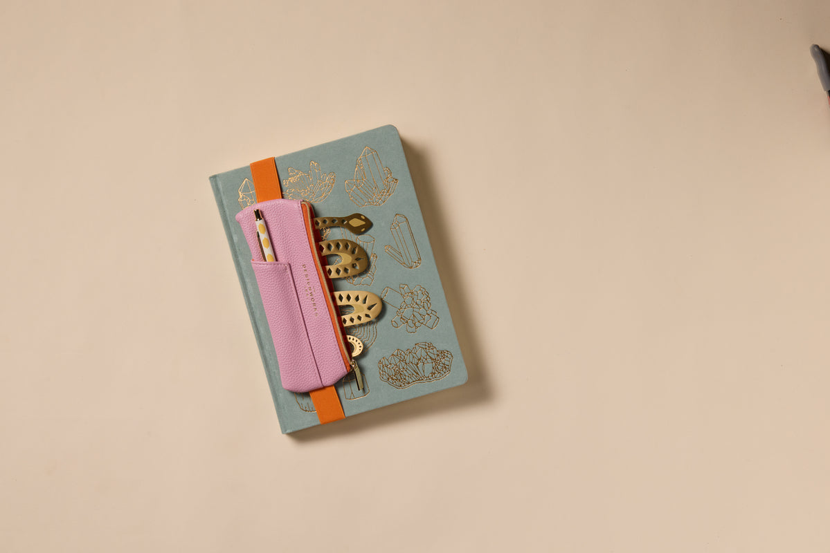 A closed journal with gold illustrations is secured by an orange band, featuring a Vegan Leather Pen Pouch in Pink + Orange attached to the cover and holding two pens. The background shows a plain beige surface.
