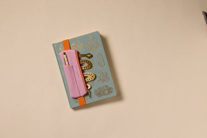 A closed journal with gold illustrations is secured by an orange band, featuring a Vegan Leather Pen Pouch in Pink + Orange attached to the cover and holding two pens. The background shows a plain beige surface.