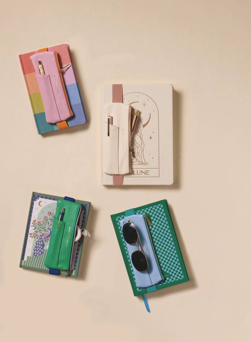 Four colorful journals, each with a pen pouch or holder attached; one features the Vegan Leather Pen Pouch in Light Blue + Green. Two pouches hold pens, one holds sunglasses, all arranged on a beige background.