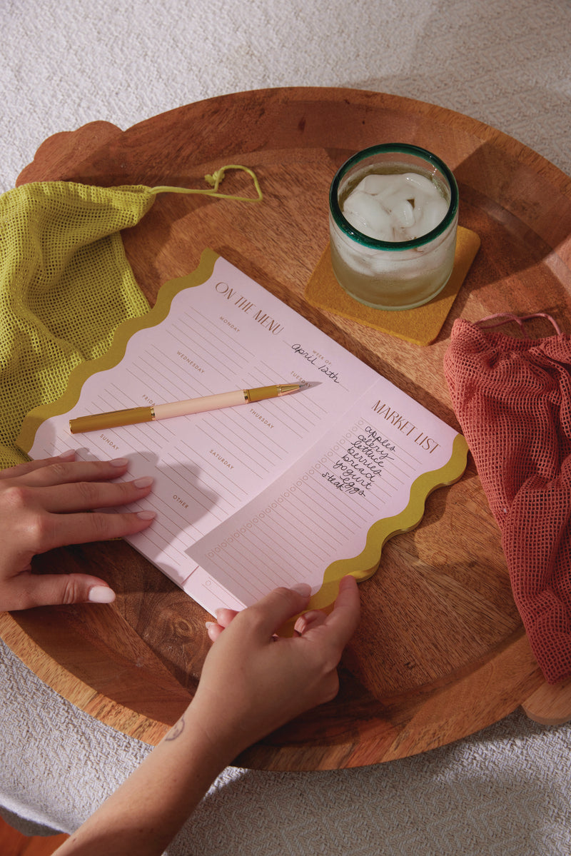Using the Meal Planner Notepad with Magnets - Pink + Chartreuse, someone writes a grocery list on its menu and market sections, placed on a wooden tray with a green pen, mesh bags, and a glass of ice water.