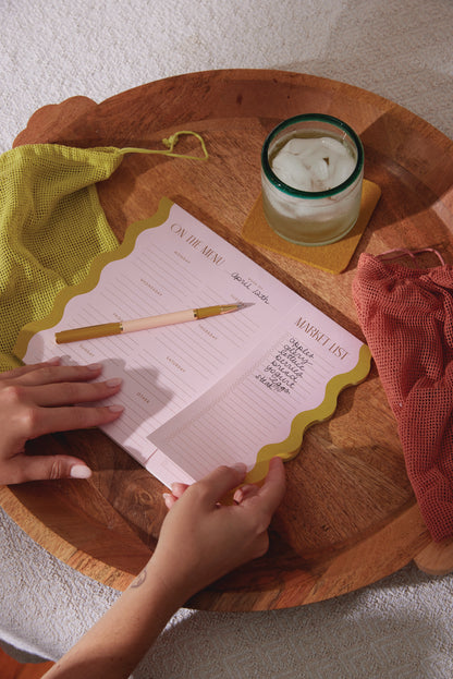 Using the Meal Planner Notepad with Magnets - Pink + Chartreuse, someone writes a grocery list on its menu and market sections, placed on a wooden tray with a green pen, mesh bags, and a glass of ice water.