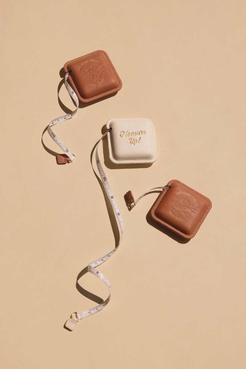 Three Pocket Tape Measure - Measure Up cases sit on a beige surface—two brown, one white with "Measure Up!" in gold. The designer tape is partially extended, looping around all three compact cases.