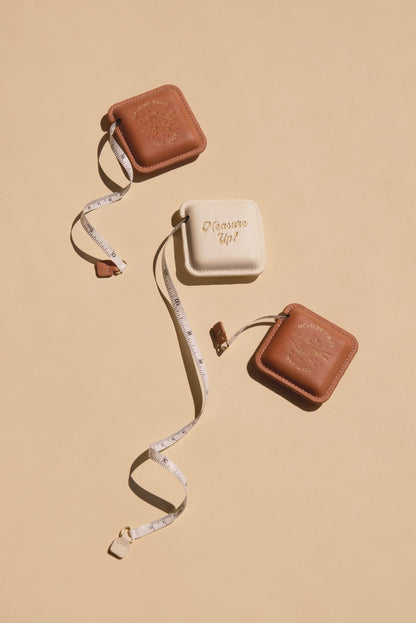 Three Pocket Tape Measure - Measure Up cases sit on a beige surface—two brown, one white with "Measure Up!" in gold. The designer tape is partially extended, looping around all three compact cases.