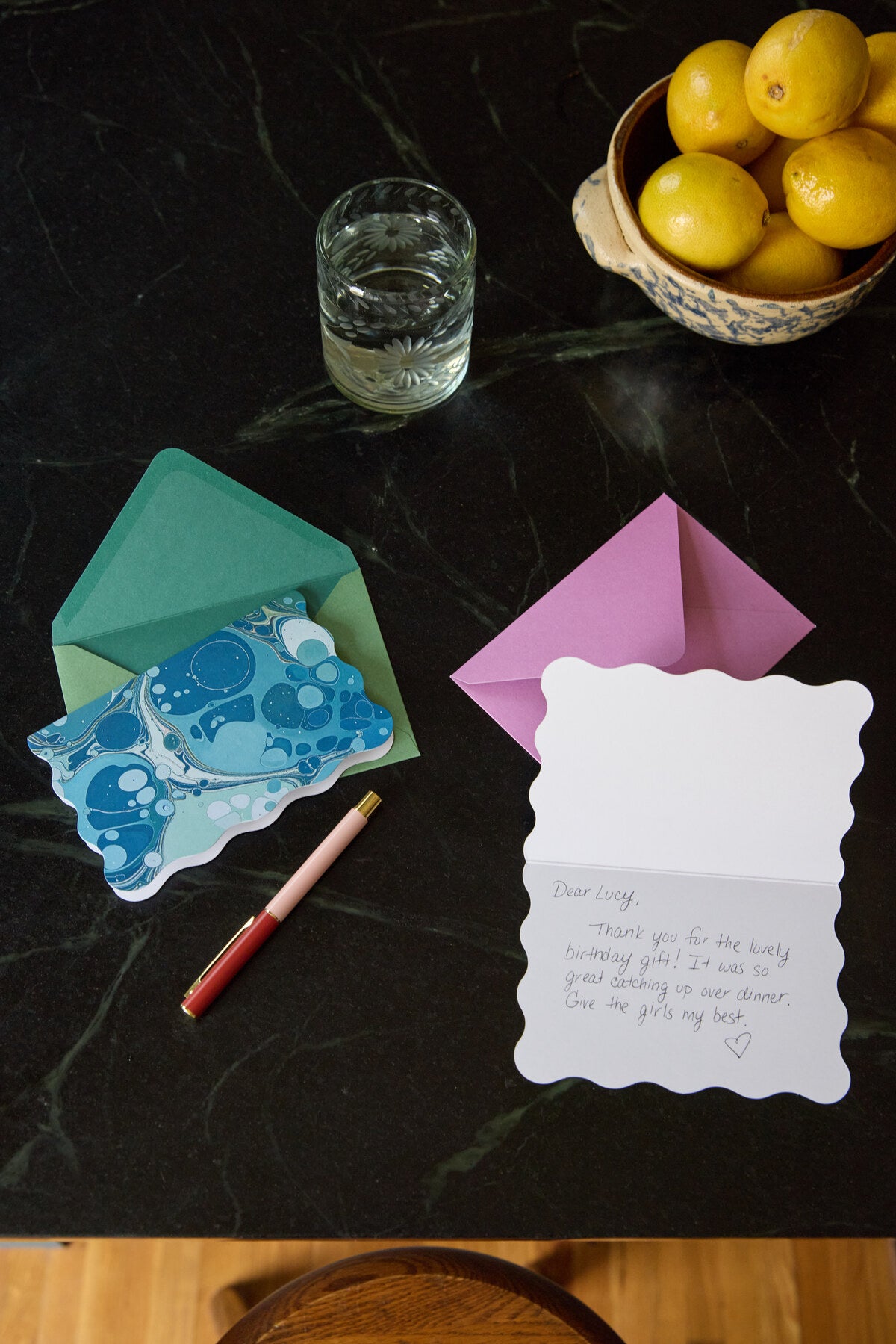 Two scalloped-edge notecards from the Boxed Marble Notecard Set - 16 Cards on a dark surface: one blue marbled card in a green envelope, and another open card with a handwritten thank-you note beside a pink pen.