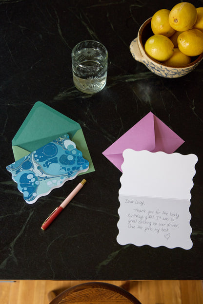 Two scalloped-edge notecards from the Boxed Marble Notecard Set - 16 Cards on a dark surface: one blue marbled card in a green envelope, and another open card with a handwritten thank-you note beside a pink pen.