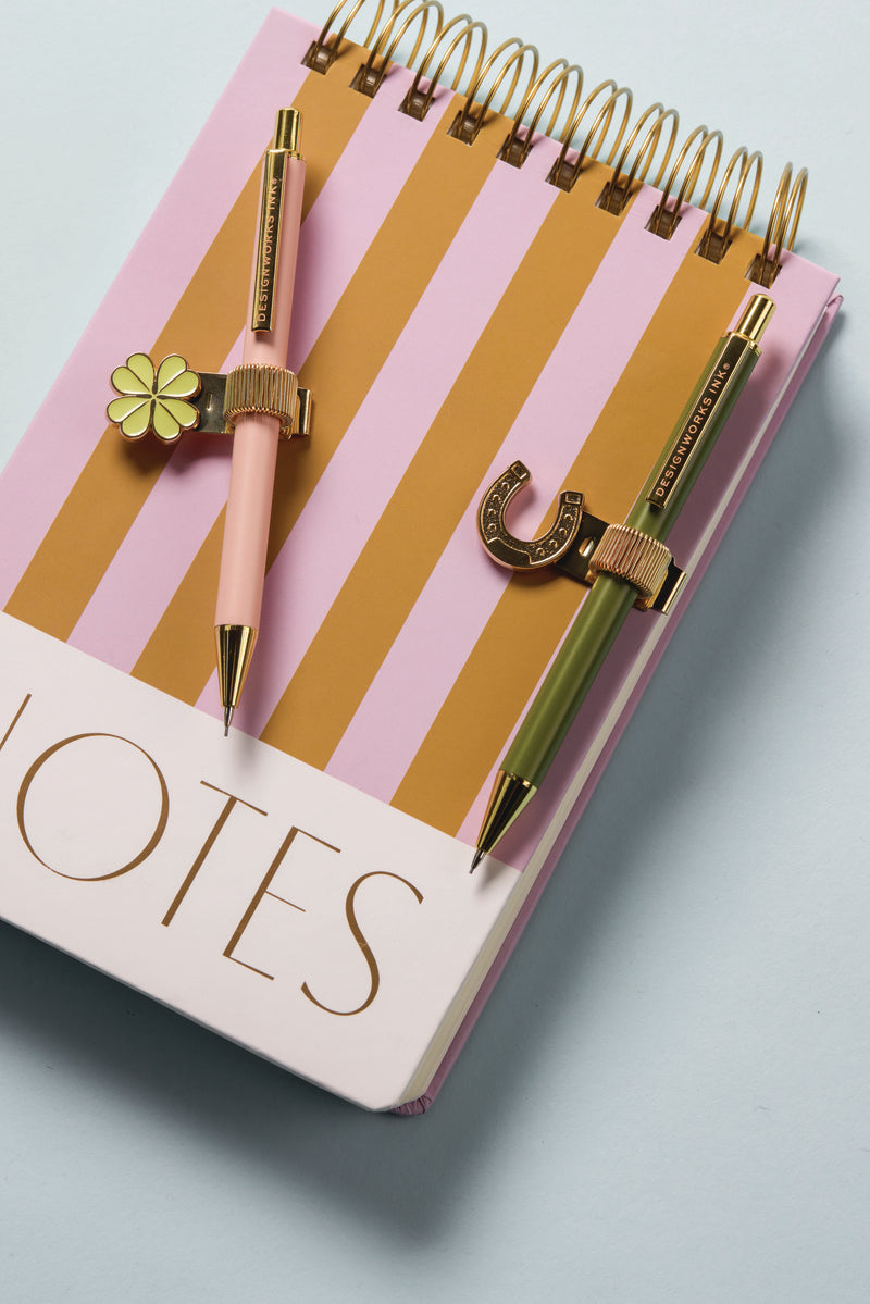 The Chunky Large Twin Wire Notepad in lilac and ochre stripes features "NOTES" on the hard cover, with two gold and pastel pens clipped to the spiral binding—one with a flower charm, the other with a cute horseshoe charm.