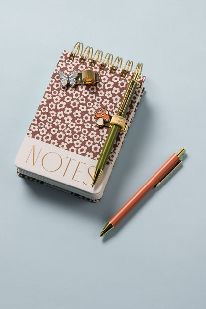 A Chunky Small Twin Wire Notepad in Burgundy Groovy Floral with 192 sheets sits on a light blue surface, labeled NOTES, topped with a green and gold pen, adorned with butterfly and heart paperclips, and an orange pen nearby.