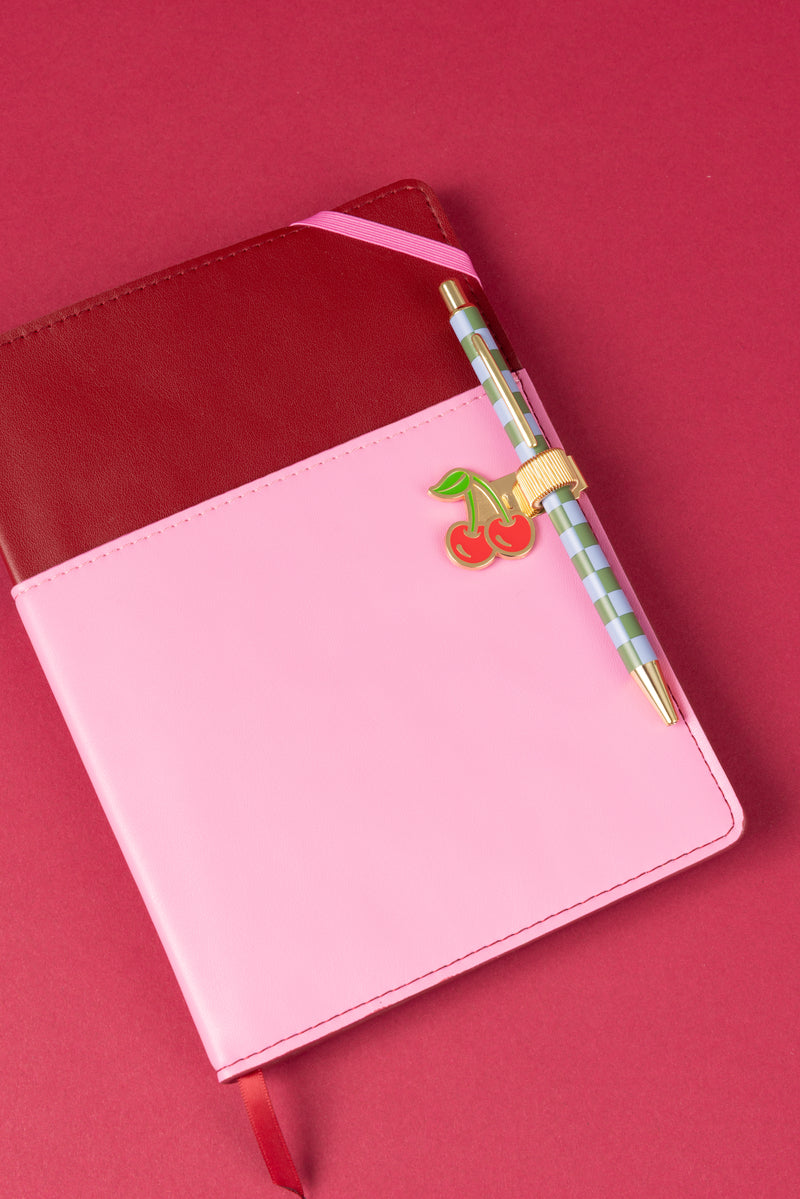 The Vegan Leather Pocket Journal - Burgundy + Pink features a maroon and pink colorblock cover with a cherry-shaped pin and comes with a striped pen with gold accents, all set against a pink background.