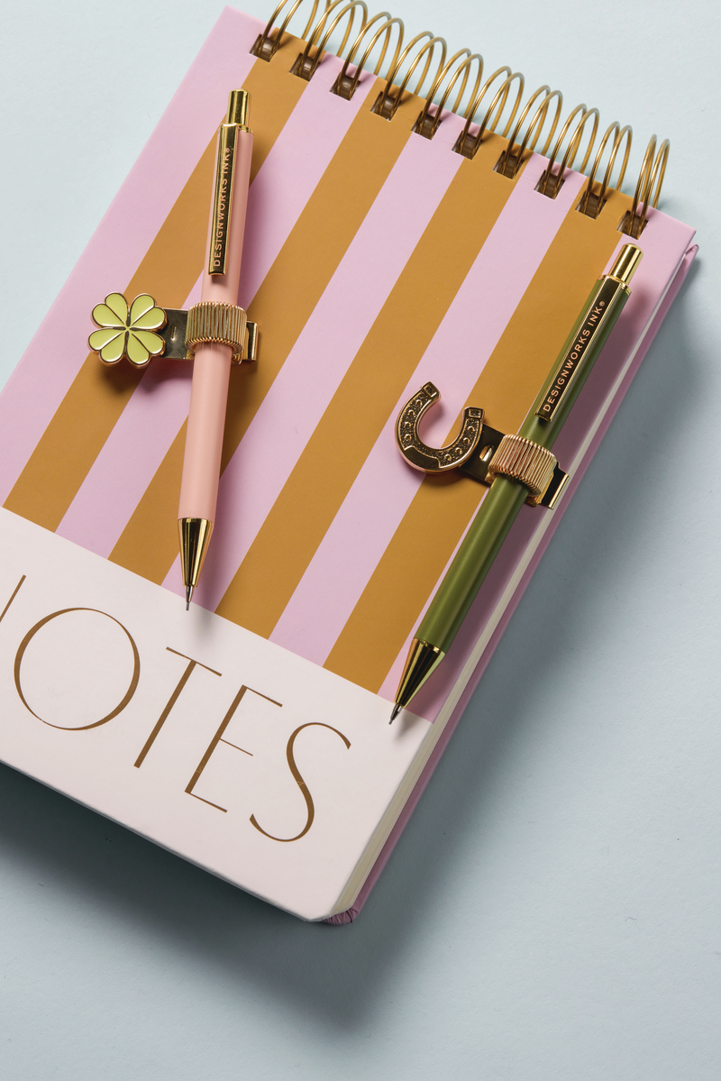 Notebook with pink and mustard stripes and two decorative pens on a light blue background