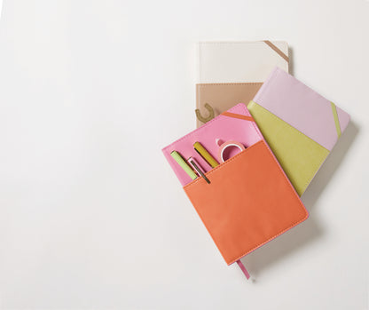 Three colorful notebooks—a Vegan Leather Pocket Journal in Pink + Chili, a white and beige notebook, and a pink journal with green accents—are arranged on a light background.