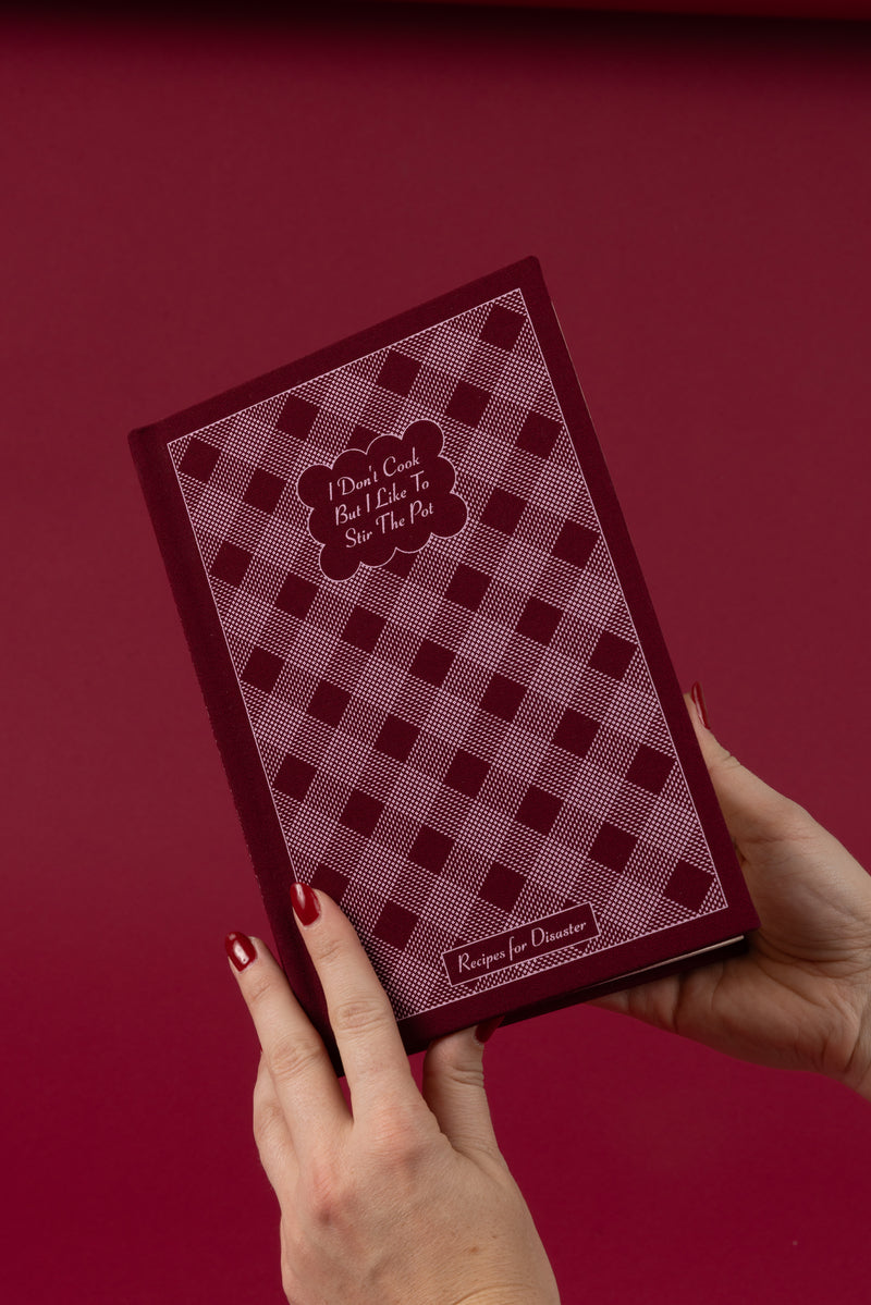 A person with red-painted nails holds the I Don't Cook Journal, featuring a maroon and pink checkered cover that reads "I Don’t Cook But I Like To Stir The Pot" and "Recipe for Disaster" on a matching maroon background.