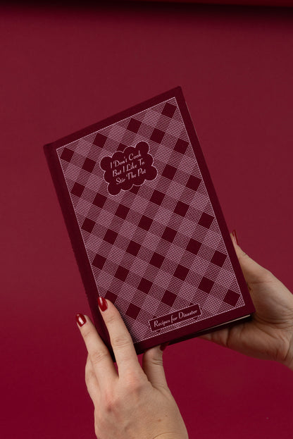 A person with red-painted nails holds the I Don't Cook Journal, featuring a maroon and pink checkered cover that reads "I Don’t Cook But I Like To Stir The Pot" and "Recipe for Disaster" on a matching maroon background.