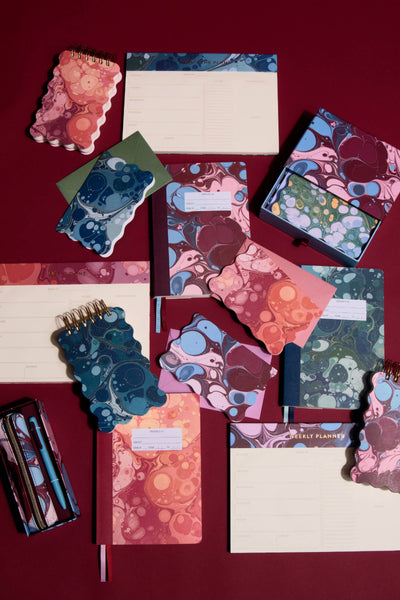 A styled flat-lay of a stationery set featuring notebooks, notepads, and a pen set with a marbled abstract pattern in shades of pink, blue, and purple, arranged on a dark red background.