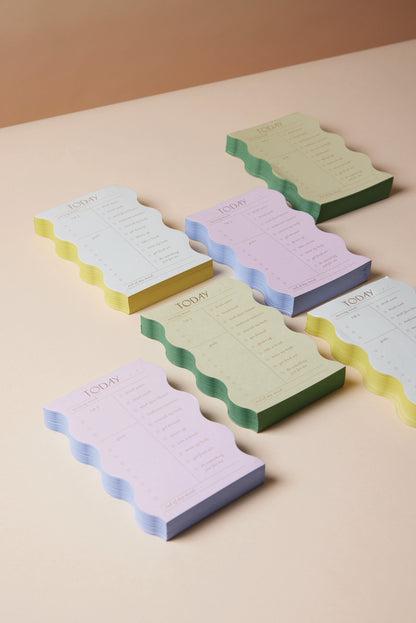 The Wavy Daily Notepad - Lilac, with its pastel lilac wavy design, is perfect for daily goals. The notepad features a "TODAY" heading and a convenient checklist, arranged on a beige surface for a stylish look.