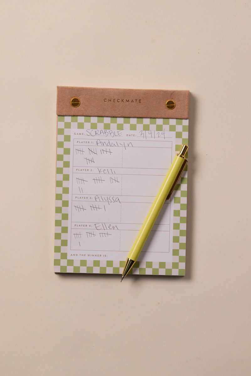The Check Mate - Game Score Post-Bound Pad features a brown header and handwritten Scrabble scores for four players, with a yellow pen on top. This 4-player score pad rests on a beige surface, ideal for organized scorekeeping.