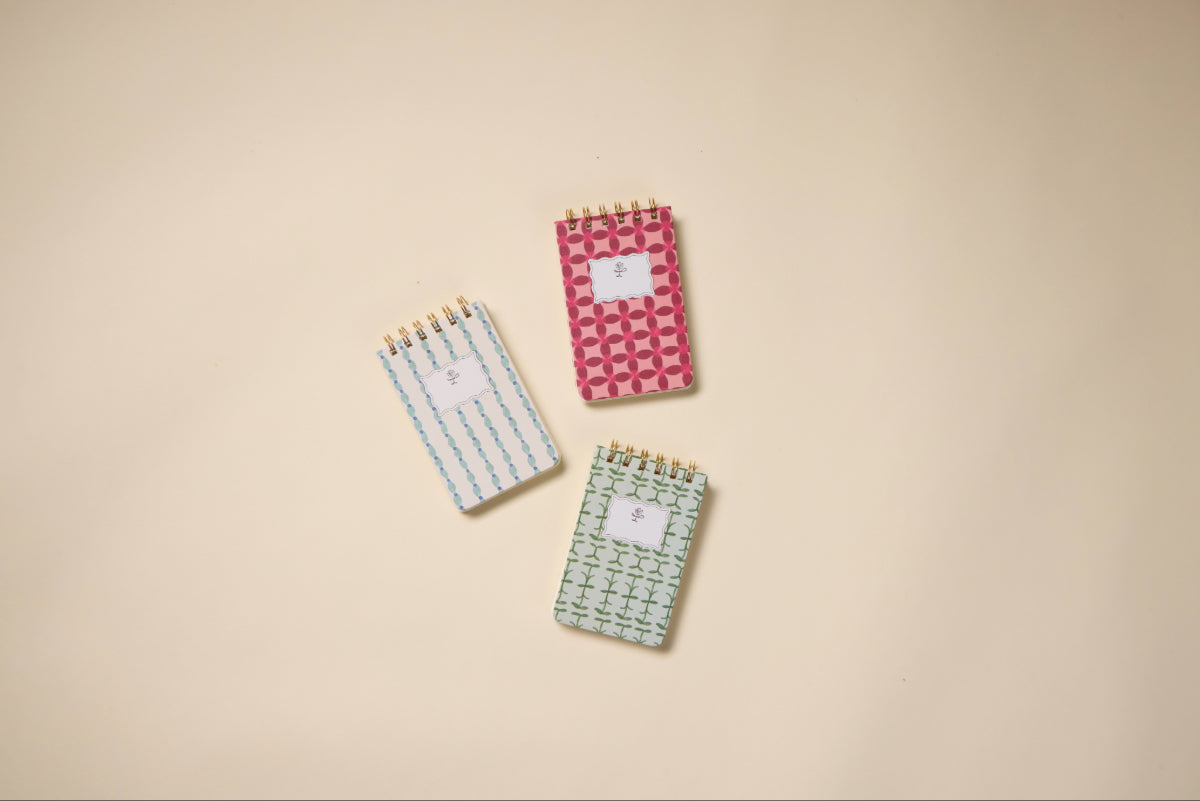 Three small spiral notepads, including a Tulip Twin Wire Notepad in Pinwheel Pink with geometric shapes, a white twin-wire notepad with lined pages, and a green one with a cross pattern, are arranged on a beige background.