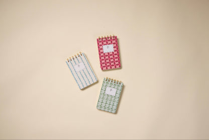 Three small spiral notepads, including a Tulip Twin Wire Notepad in Pinwheel Pink with geometric shapes, a white twin-wire notepad with lined pages, and a green one with a cross pattern, are arranged on a beige background.