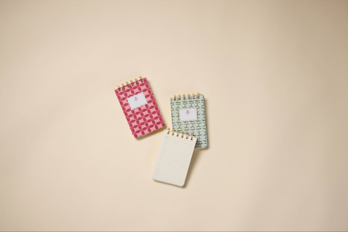 Three small notepads are arranged on a beige surface: one has a Pinwheel Pink cover like the Tulip Twin Wire Notepad, another has a green patterned cover, and the third, a Twin-Wire Notepad, is open to reveal lined pages.