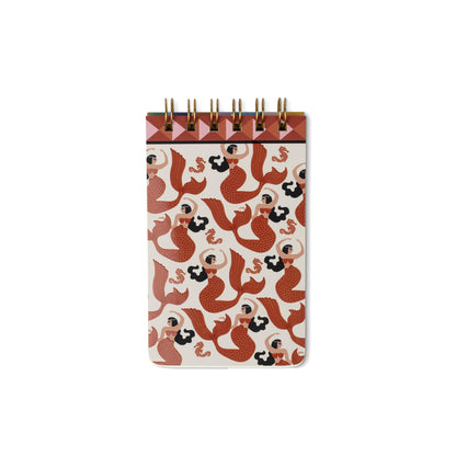 A small spiral-bound notebook with a cream cover decorated with a repeating pattern of red and black mermaids, featuring a brown top band and gold wire spirals, shown on a plain white background.