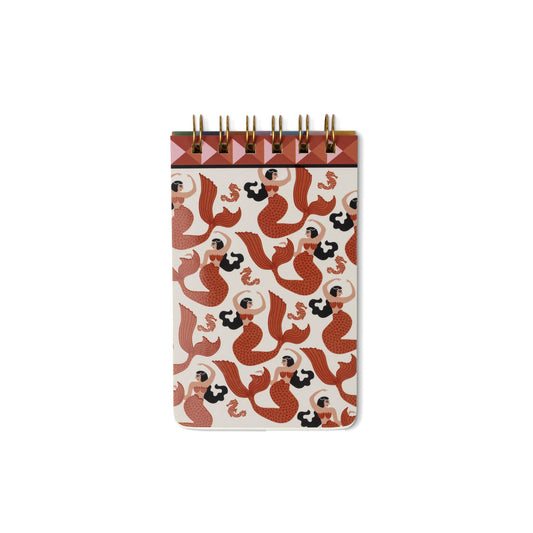 A small spiral-bound notebook with a cream cover decorated with a repeating pattern of red and black mermaids, featuring a brown top band and gold wire spirals, shown on a plain white background.