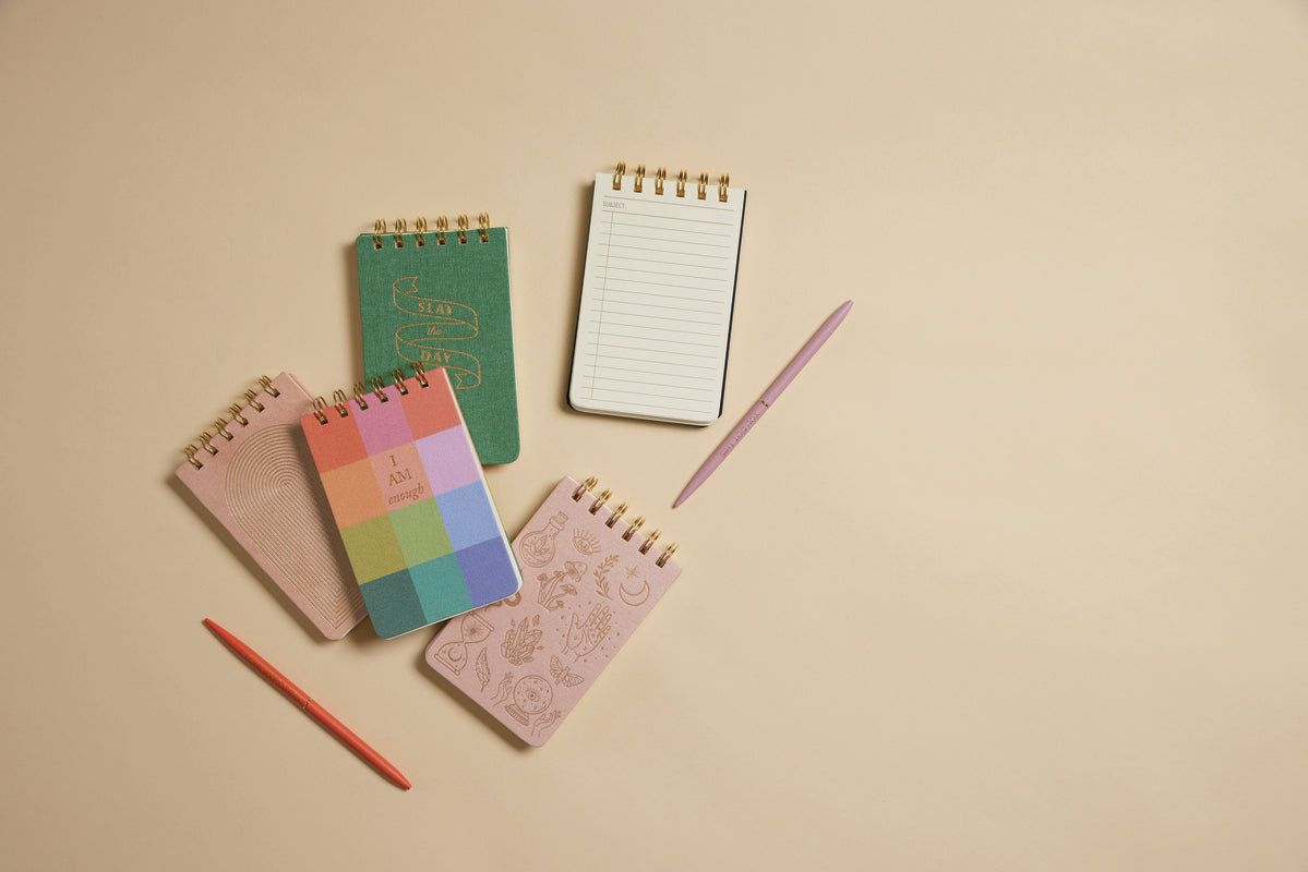 Five Twin Wire Notepad - Rainbow Check notepads, each with a unique colorful cover, are arranged on a beige surface with a red pencil and pink pen. One open portable notepad reveals its lined pages.