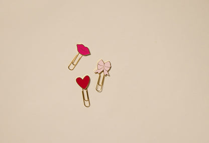 The Set of 3 Paperclips - Love features cute designs—a pink lips clip, a pink bow clip, and a red heart clip—on a beige background, making charming office supplies to brighten any workspace.
