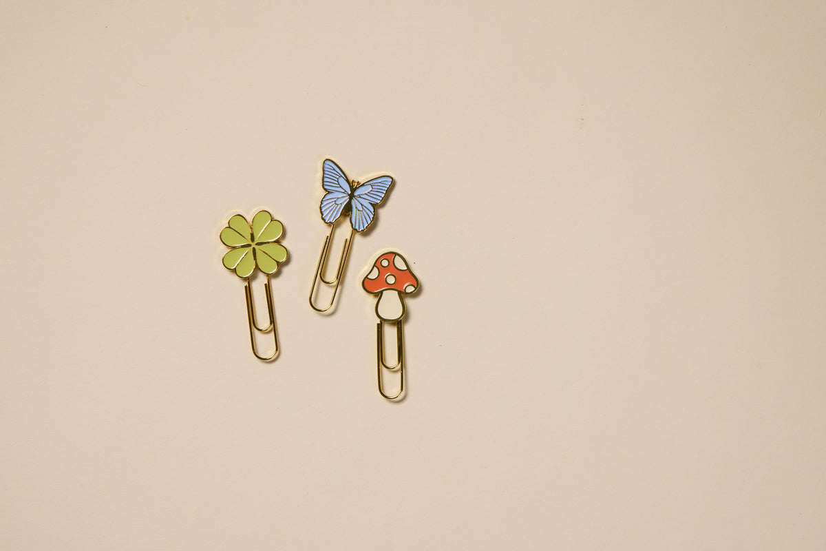 The Set of 3 Paperclips - Nature includes gold paperclips with playful tops: a green four-leaf clover, blue butterfly, and red mushroom—charming planner accessories displayed on a plain beige background.