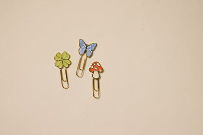 The Set of 3 Paperclips - Nature includes gold paperclips with playful tops: a green four-leaf clover, blue butterfly, and red mushroom—charming planner accessories displayed on a plain beige background.